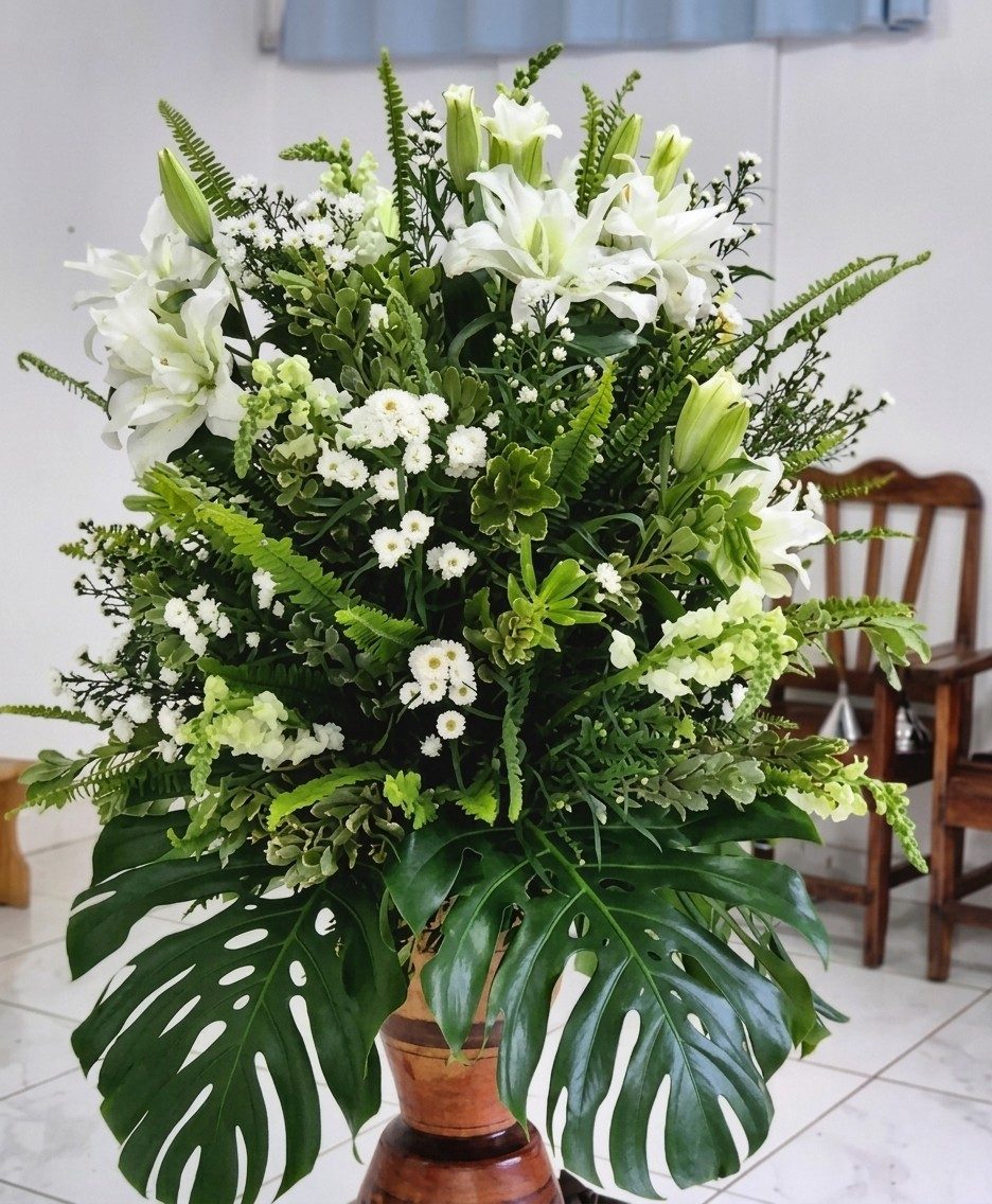 Arranjo alto em tons de branco e verde, com lírios, crisântemos, bocas-de-leão, samambaias e folhas de costela-de-adão, sobre um pedestal de madeira.