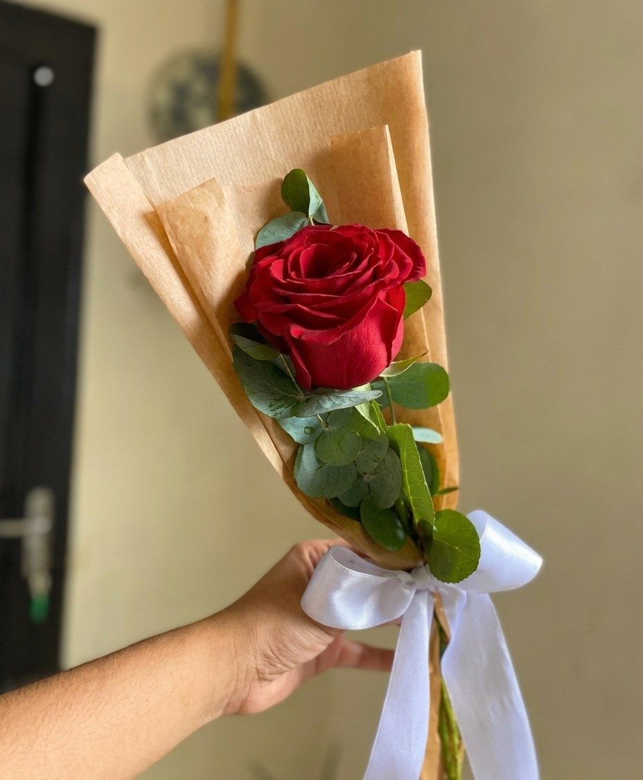 Uma rosa vermelha com folhas de eucalipto, envolta em papel kraft e amarrada com laço de cetim branco.