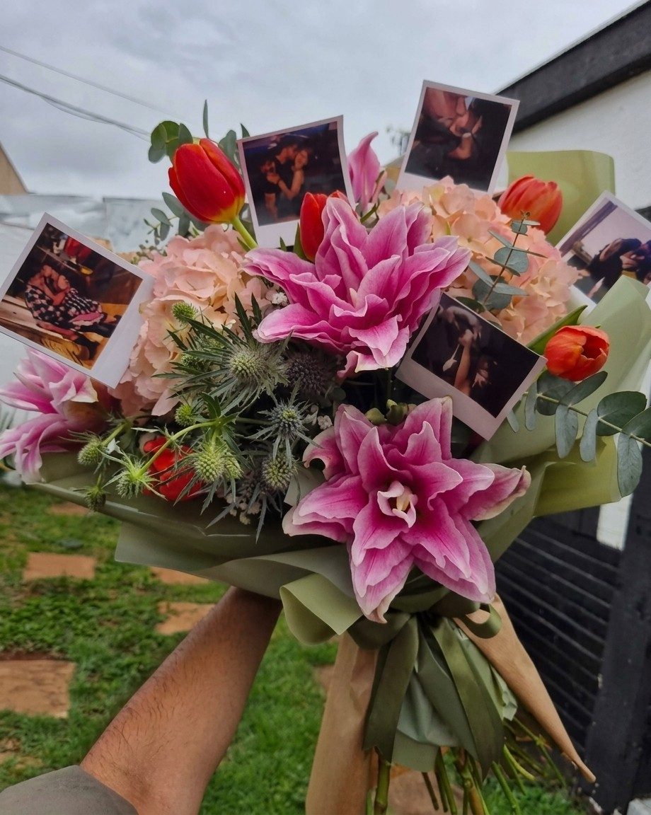 Buquê com lírios rosa, tulipas vermelhas, hortênsias cor de pêssego, cardo e eucalipto, decorado com fotos instantâneas de um casal e embrulhado em papel verde.