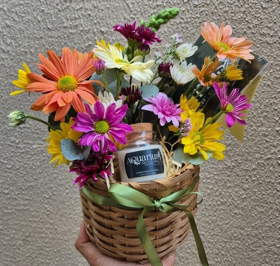 Cesta de palha com crisântemos coloridos em laranja, amarelo, pink e branco, uma vela artesanal do Aquarius e um cartão, amarrada com fita verde e segurada na mão.