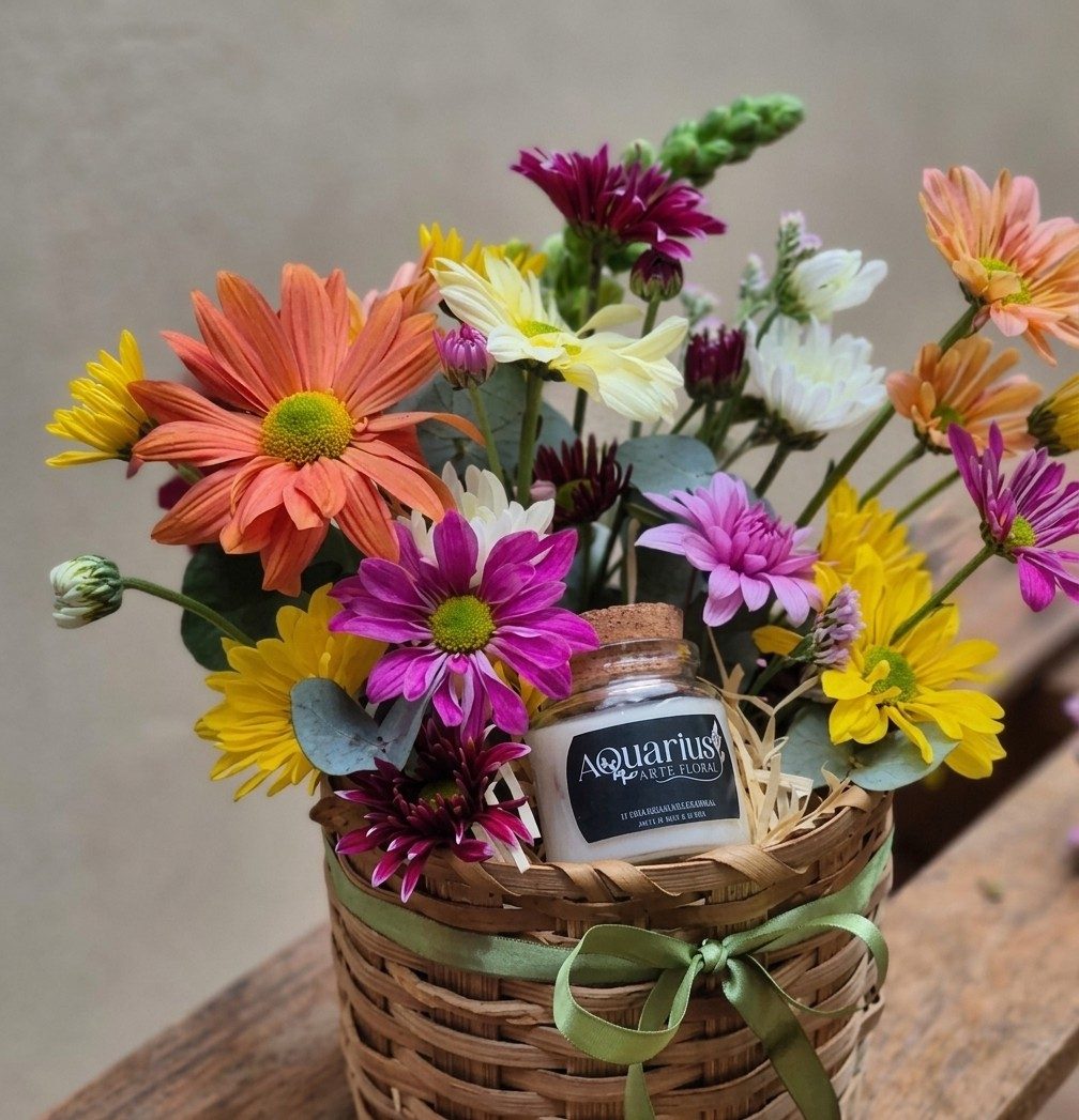 Cesta de palha com crisântemos coloridos em laranja, amarelo, pink e branco e uma vela artesanal do Aquarius, amarrada com fita verde.