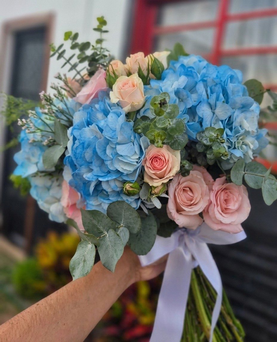 Buquê de noiva com hortênsias azuis, rosas cor de pêssego e eucalipto, amarrado com fita lilás.