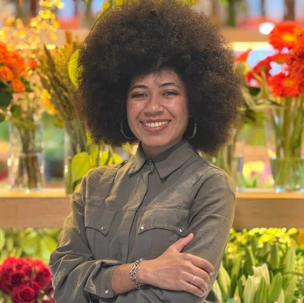 Mariana Santos sorrindo, de braços cruzados, em frente a prateleiras com vasos de flores.
