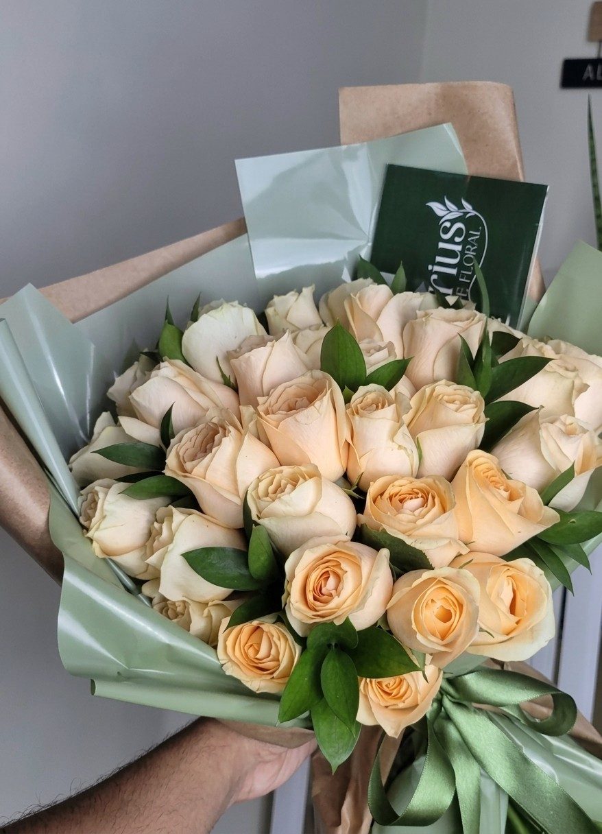 Buquê de rosas champanhe com folhas de rúscus, envolto em papel verde-sálvia e kraft, com cartão do Aquarius e fita de cetim verde.